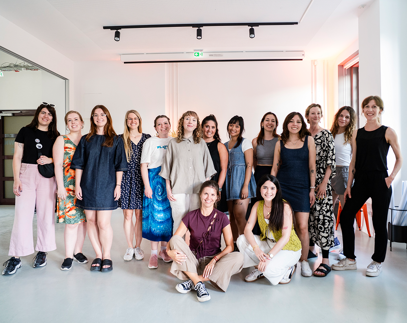 Group of fifteen women posing and smiling in a bright indoor room with white walls and modern lighting.